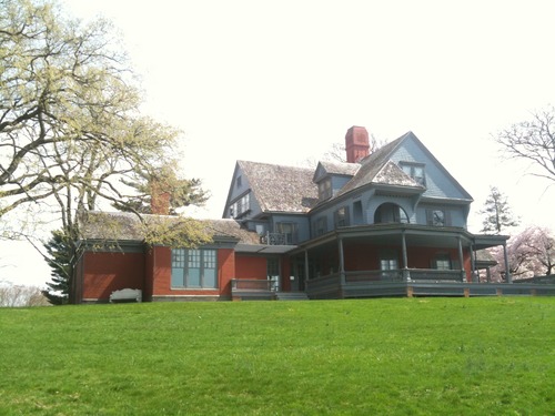 A two-story Colonial Revival style house with a wrap-around porch stands atop of grassy rise.