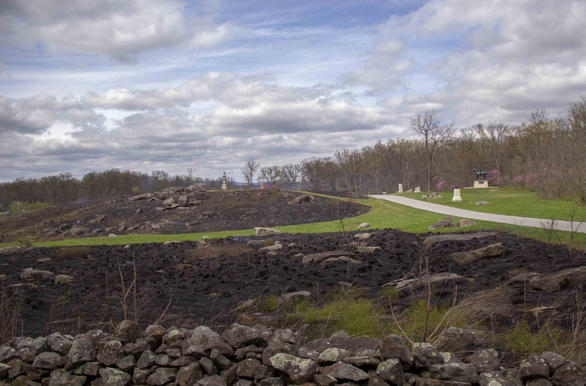 A low stone wall, then a boulder strewn blackened area, then a strip of green grass, then another boulder strewn blackened area in the distance. Along the right is a road with another section of green grass and multiple monuments in front of a tree line.