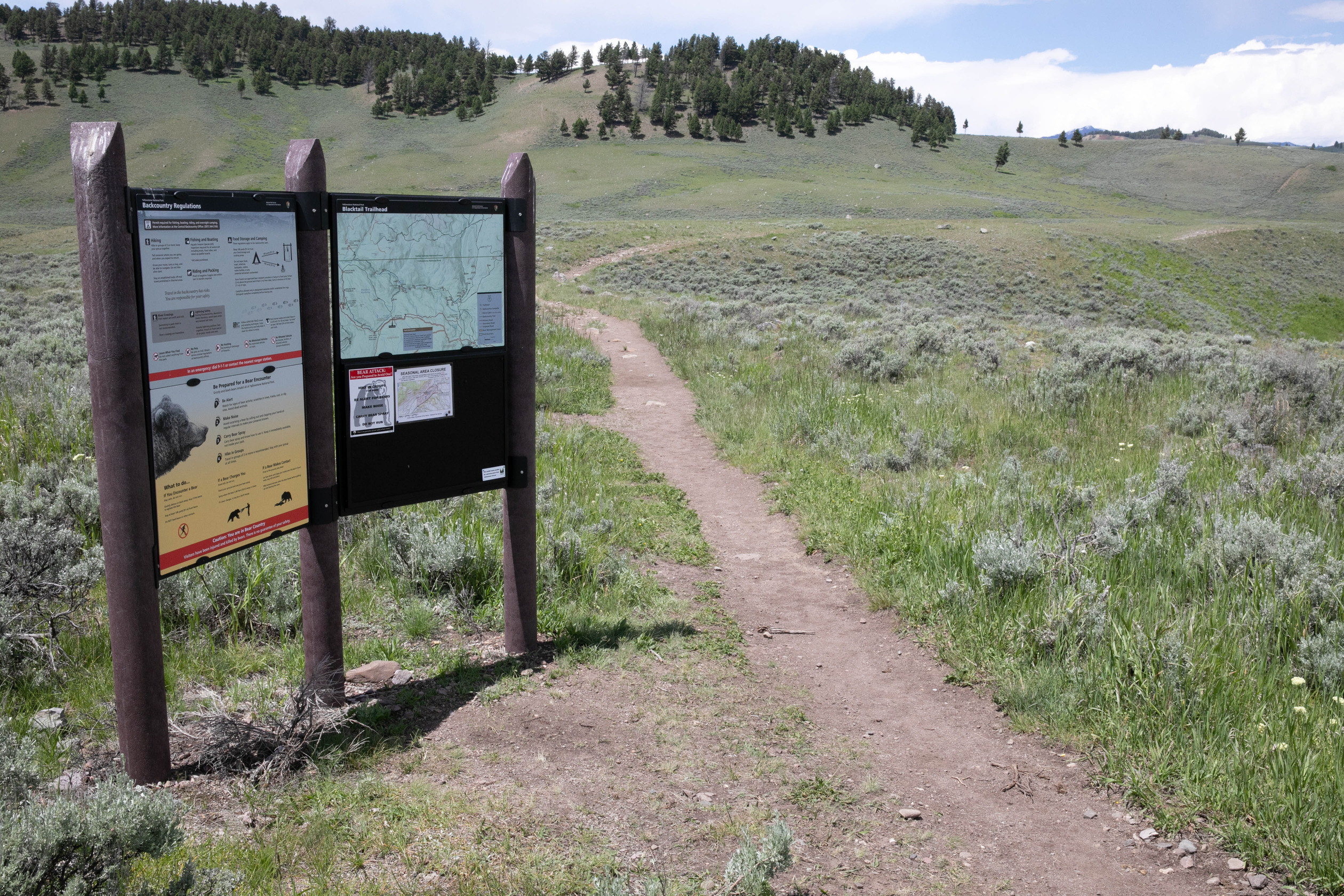 A trail passes a sign on the right and continues out into sagebrush covered terrain.