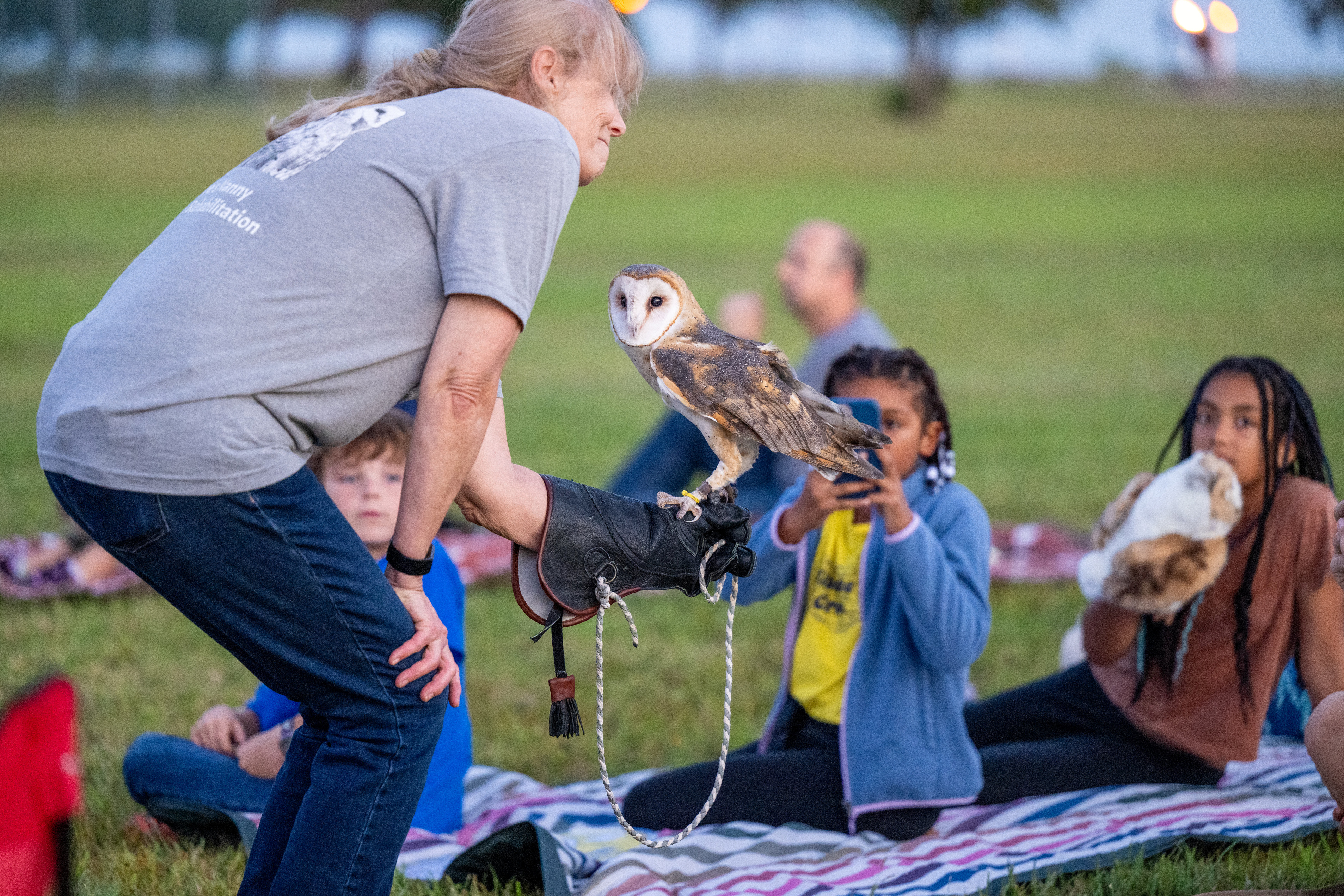 A group of children and adults sitting on a grass watch a woman holding an owl.