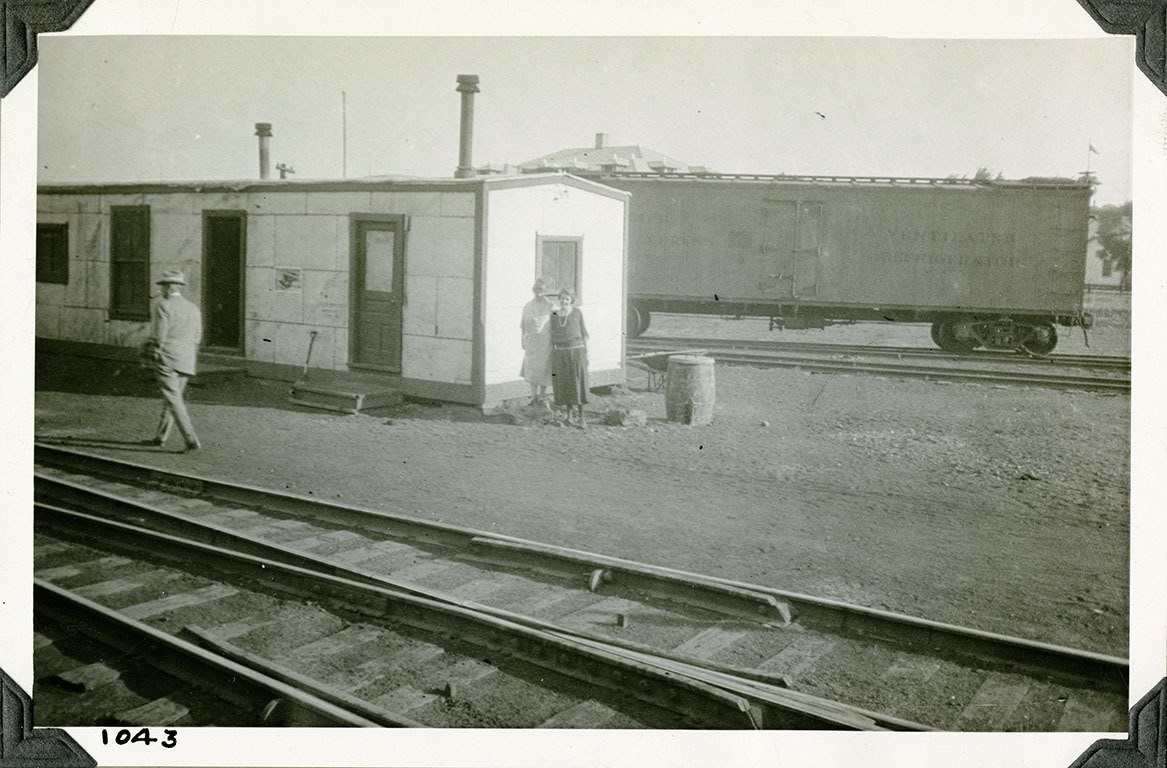 This is an historic black and white photograph from the Scotty's Castle Historic Photograph Collection, Death Valley National Park of two woman standing behind railroad track where trains can switch track in front of white shed with dark trim. Man walking in front of shed to left. Box car on tracks in background. Number in black ink in lower left corner.