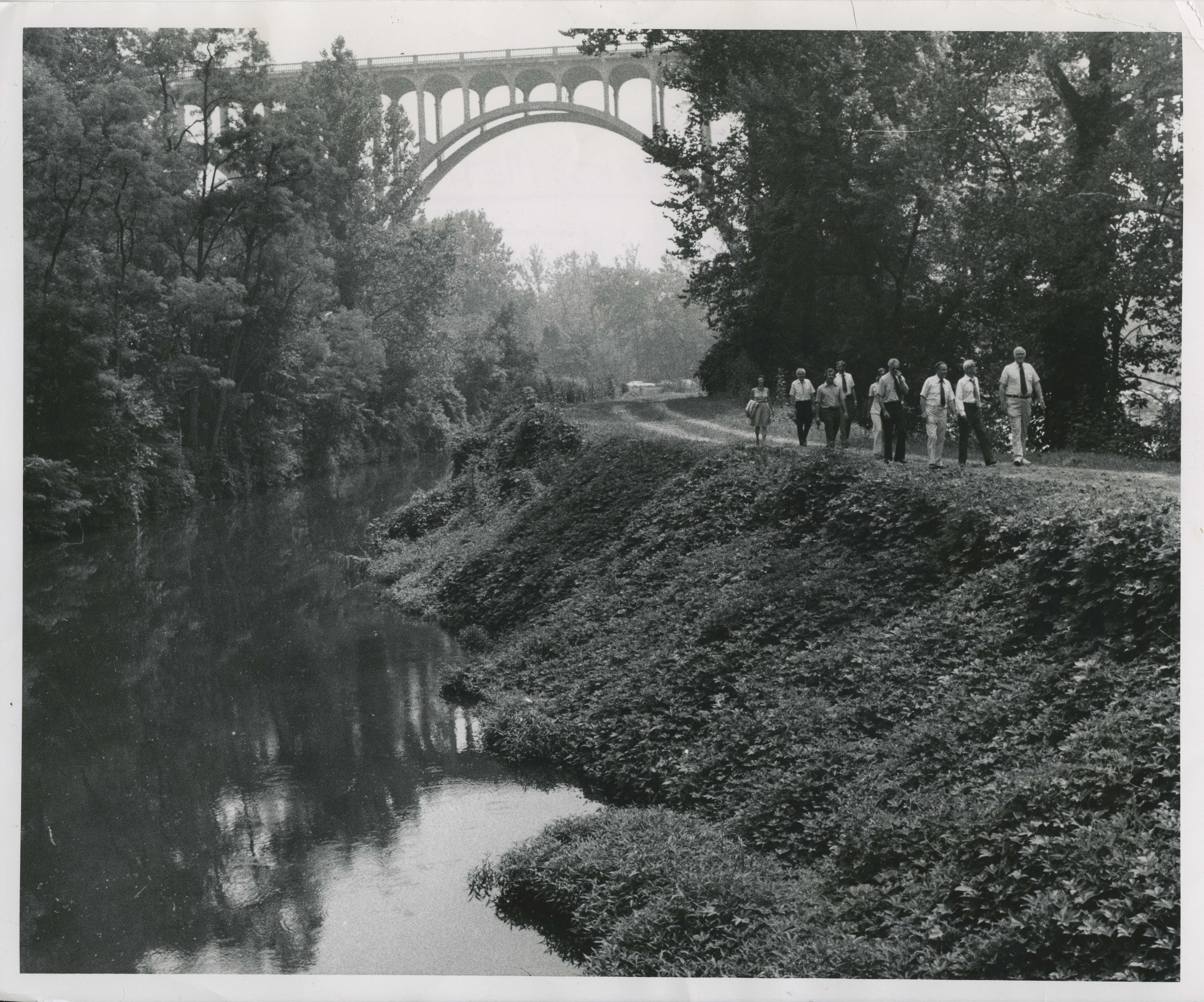 Black and white photo of ten people in shirts and ties walking along a wooded path next to a body of water with a high arched bridge overhead in the background.