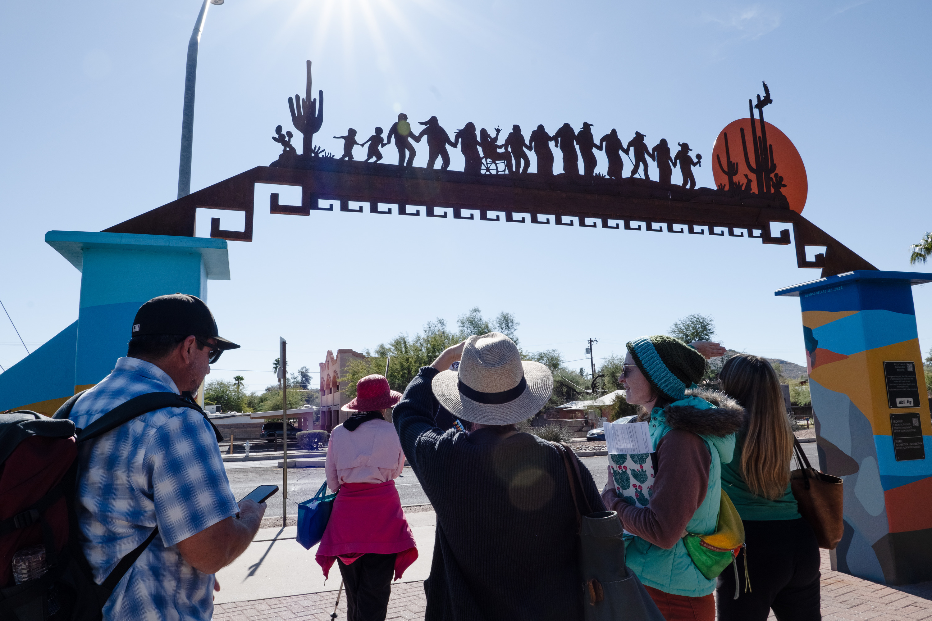 Five people face towards the sun to view a metal archway that shows saguaros and a chain of people of varying abilities holding hands and walking towards a sun