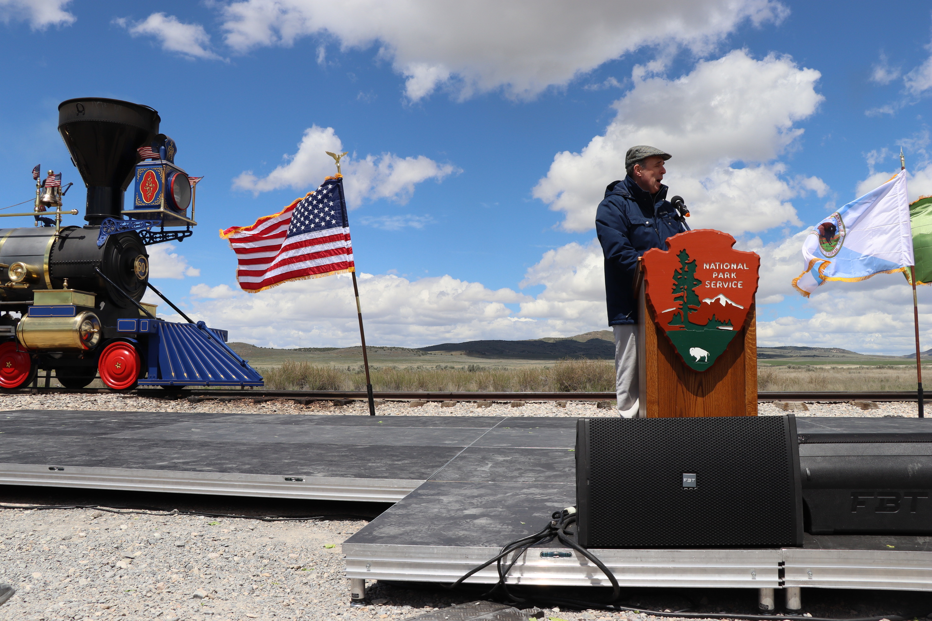 A man in black suit and grey newsboy cap talks into a microphone in front of railroad tracks, a blue sky, and three flags in the background with bright blue skies.
