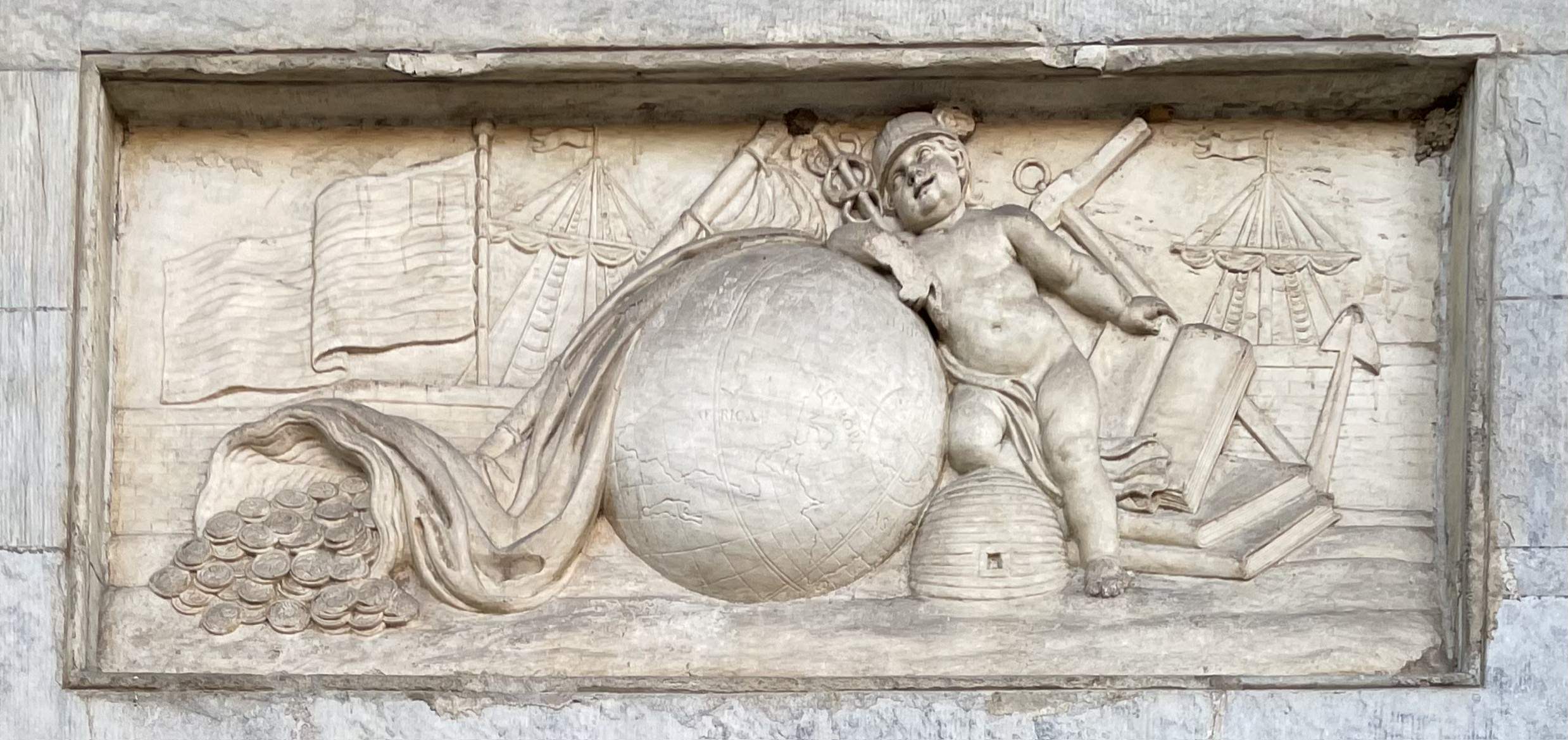 Stone carving with pictorial representations of the early economy: a globe, a baby, a beehive, a stack of books, ships, and coins spilling from a cornucopia.   