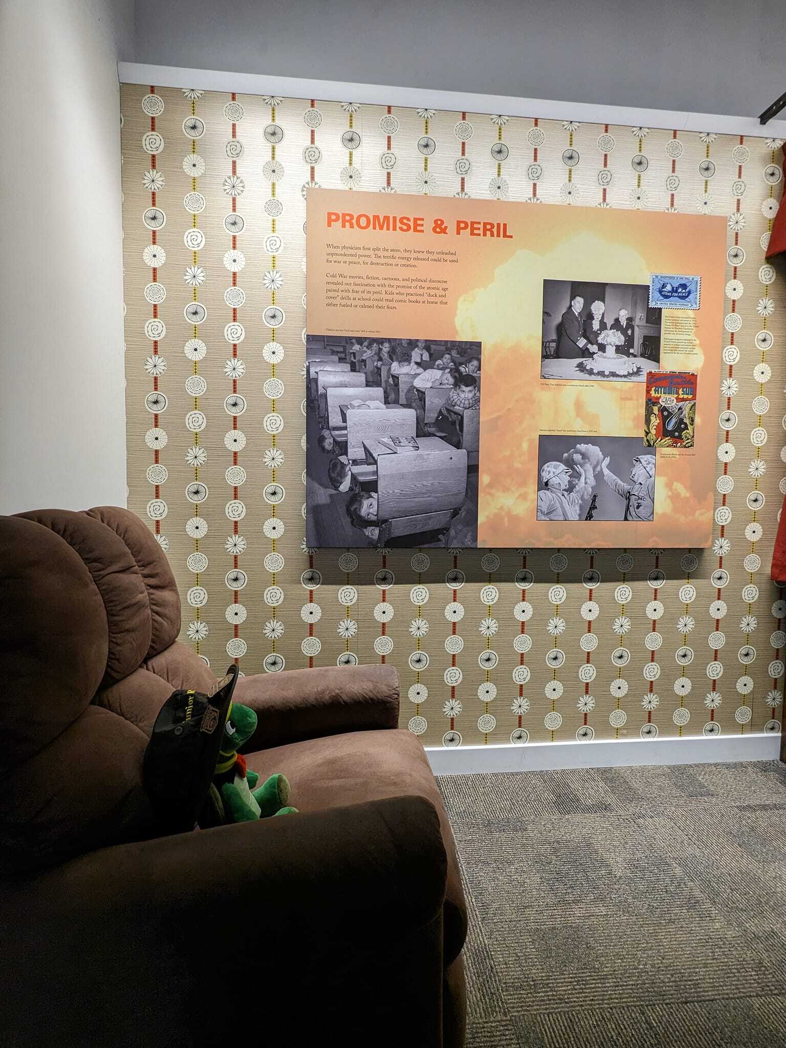 Plush turtle sitting in an armchair wearing a green bucket hat. On the wall next to him, a large panel labeled "Promise & Peril" with a few pictures from life during the Cold War
