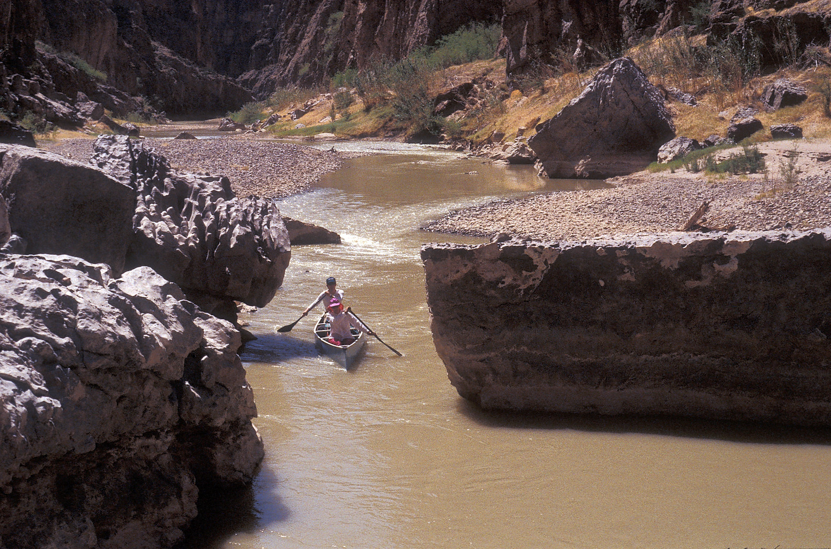 The photo is taken from above, looking down on a river and two canoeists navigating a narrow channel between two large boulders that block the rest of the river.