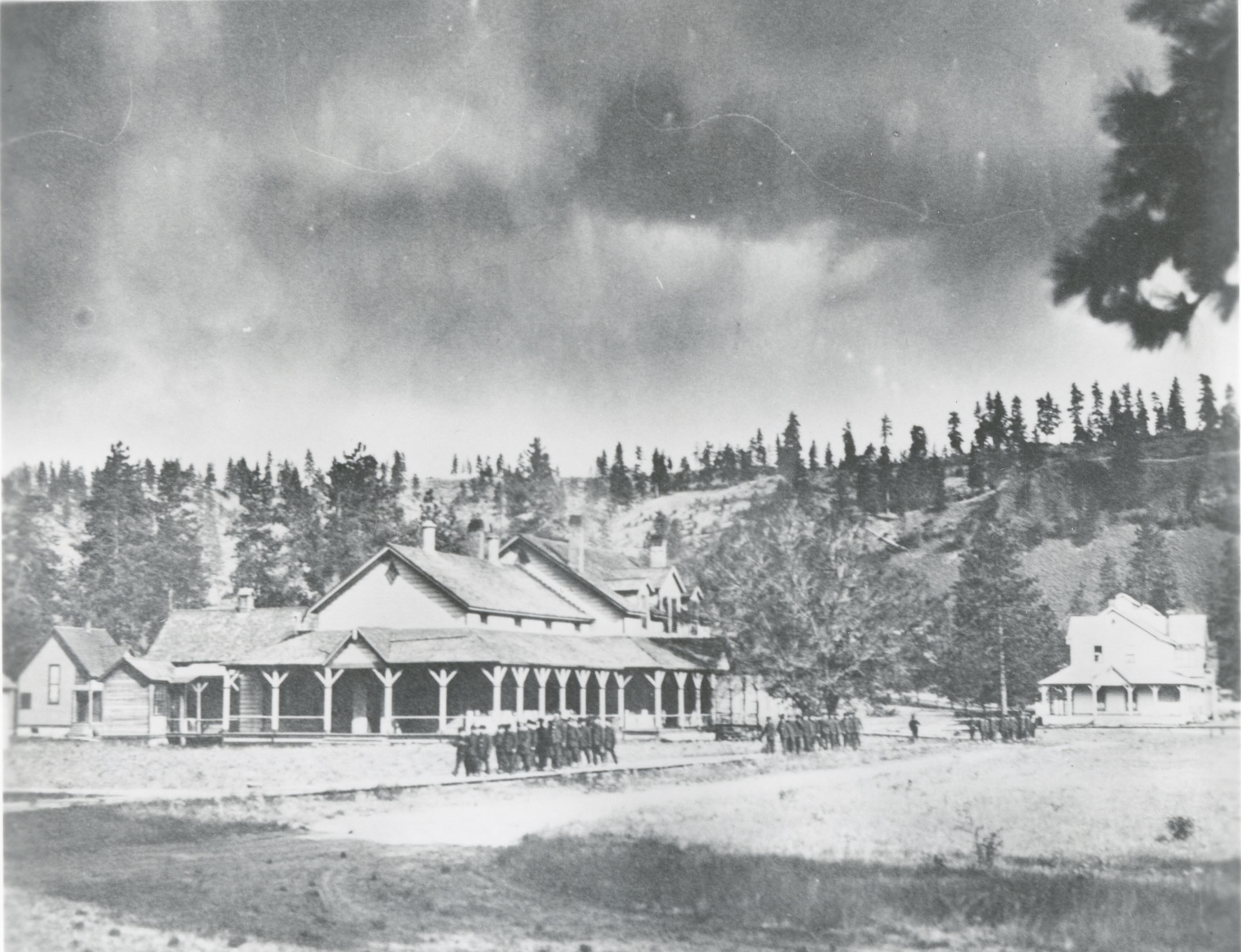 Faded black and white photograph of children walking in rows along a boardwalk in front of a large building with wrap around porch.