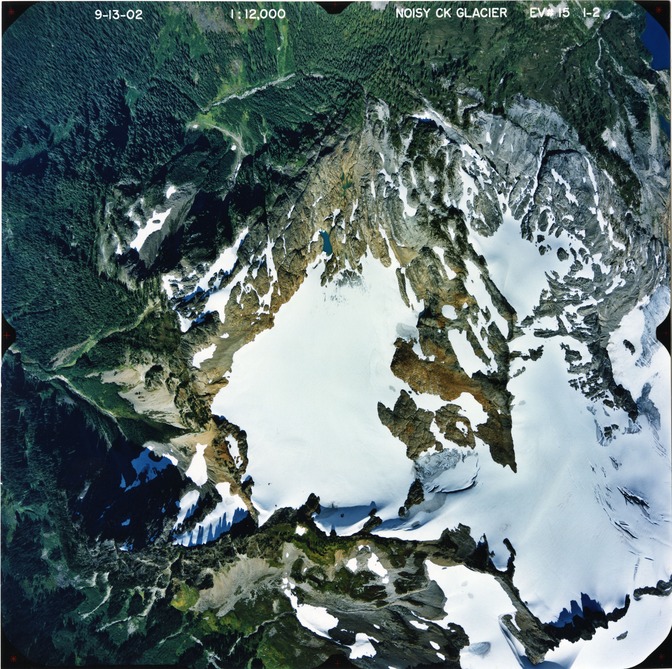 Color aerial photograph of glacier on mountain peak, surrounded by forested valleys.