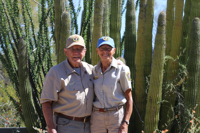 An older couple wearing ball caps and grey shirts with NPS Volunteer logos stand in front of an organ pipe cactus.