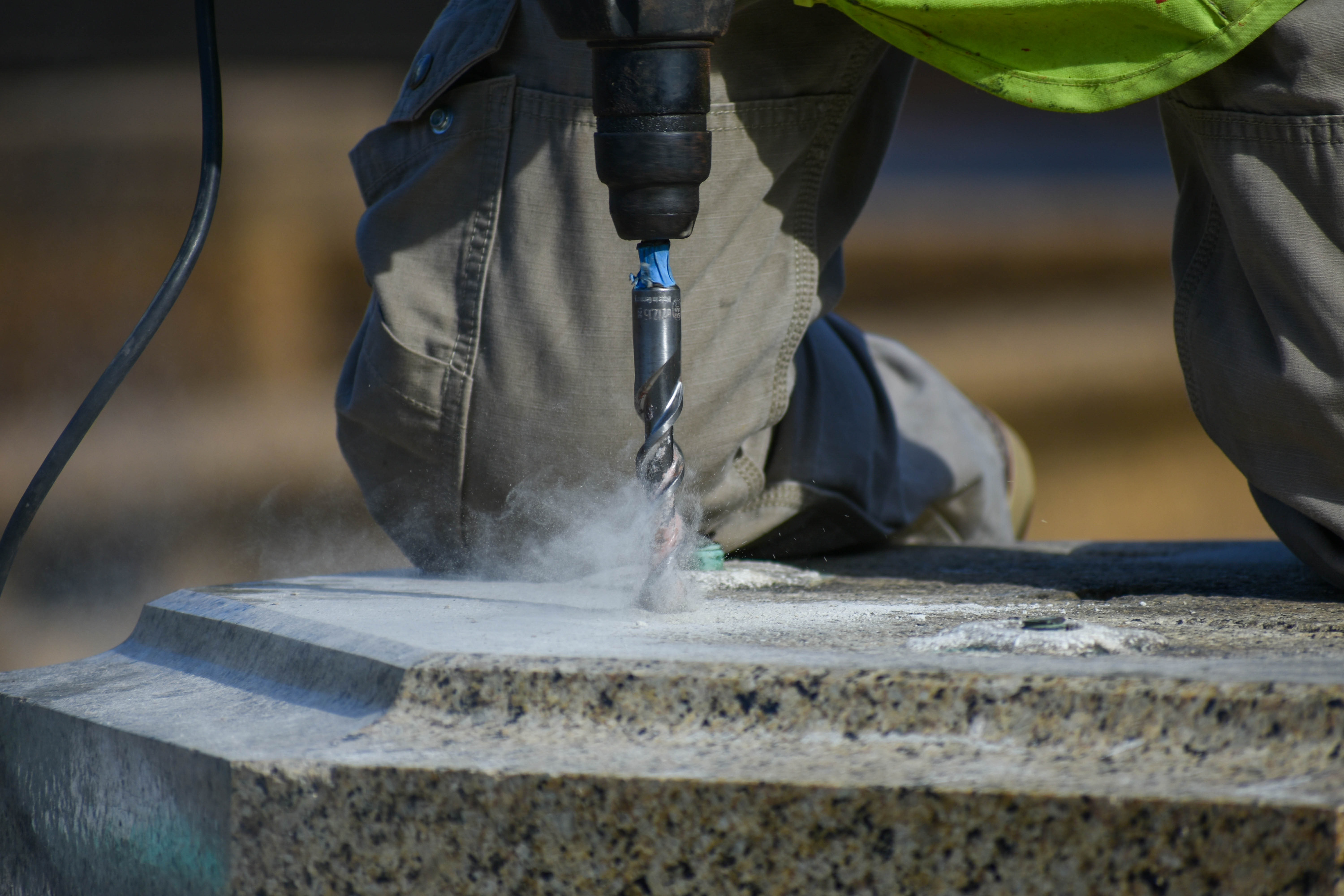 A close-up photo of a man on his knees drilling into a granite platform. Fine particles fly up from the drill and clouds of dust surround it.