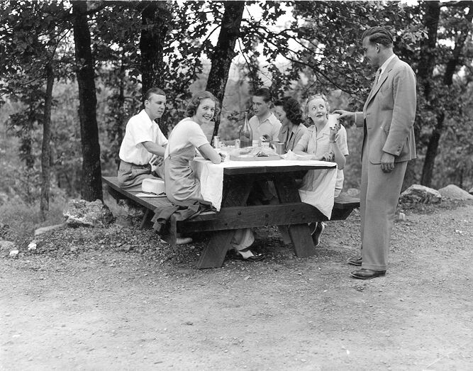 A family of 5 sit at a picnic table. They have a white tablecloth strewn across the table with dishes and bottles on top. A man stands next to them handing a woman a cup.