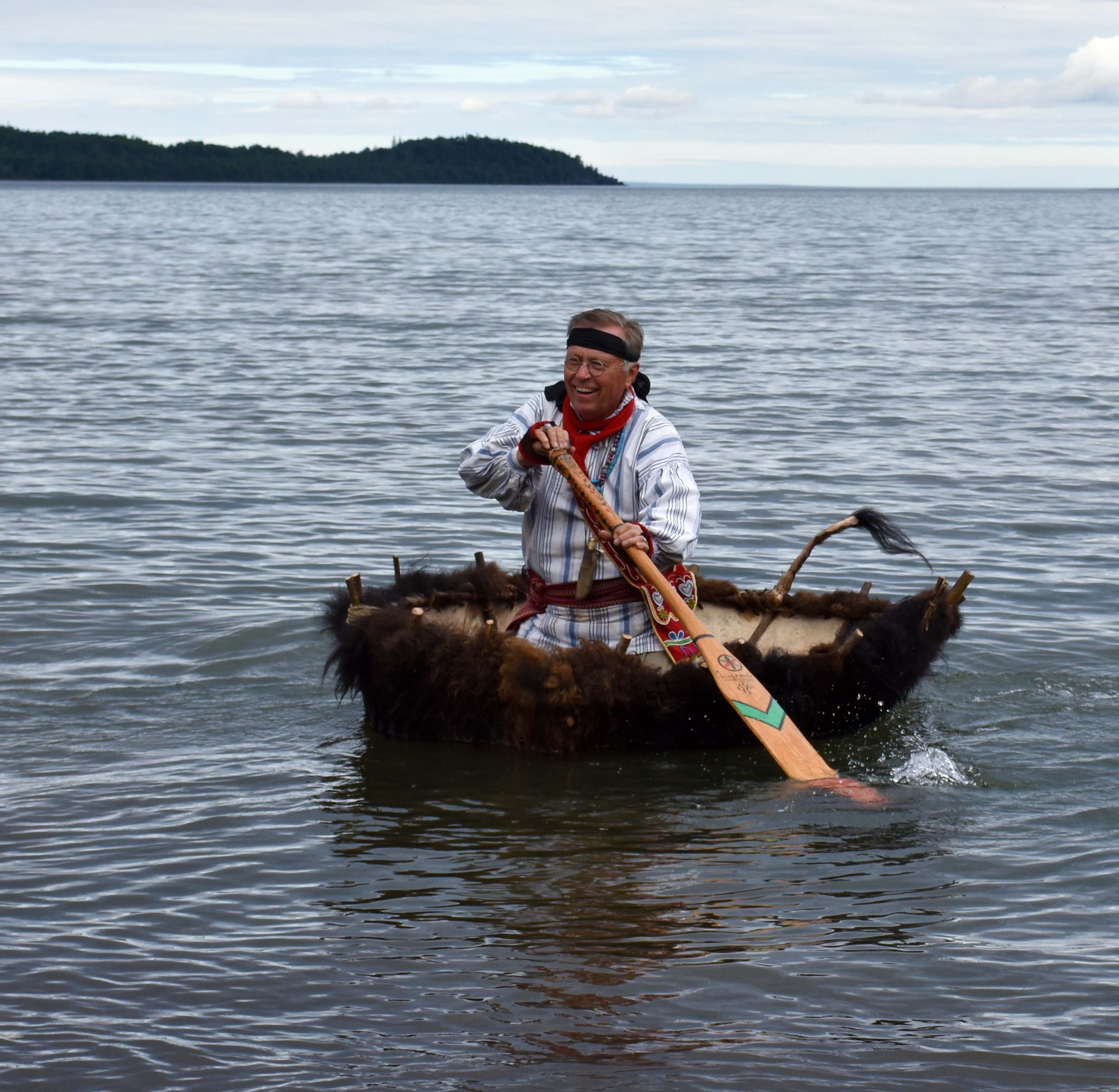A person kneeling in a small boat made of bison hide holds a decorated wood paddle. The boat is floating in gentle waves in a bay.