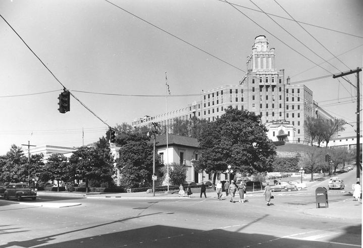 Glossy black & white photographic print, street scene; Photography of south end of Bathhouse Row and intersection of Reserve & Central Avenues; shows flag pole with flag flying in corner lawn of administration building, Lamar Bathhouse flag and antennae, south end of Buckstaff Bathhouse; Army-Navy Hospital with sign still in place over entrance, aerial walkway between hospital and Eastman Hotel annex; concrete boulevard in Central, telephone poles by Bathhouse Row; people passing on sidewalk along Bathhouse Row hedges