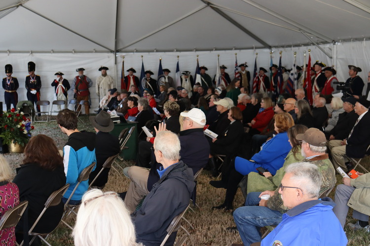 Hundreds of people sit in chairs or stand along the back of a tent. 