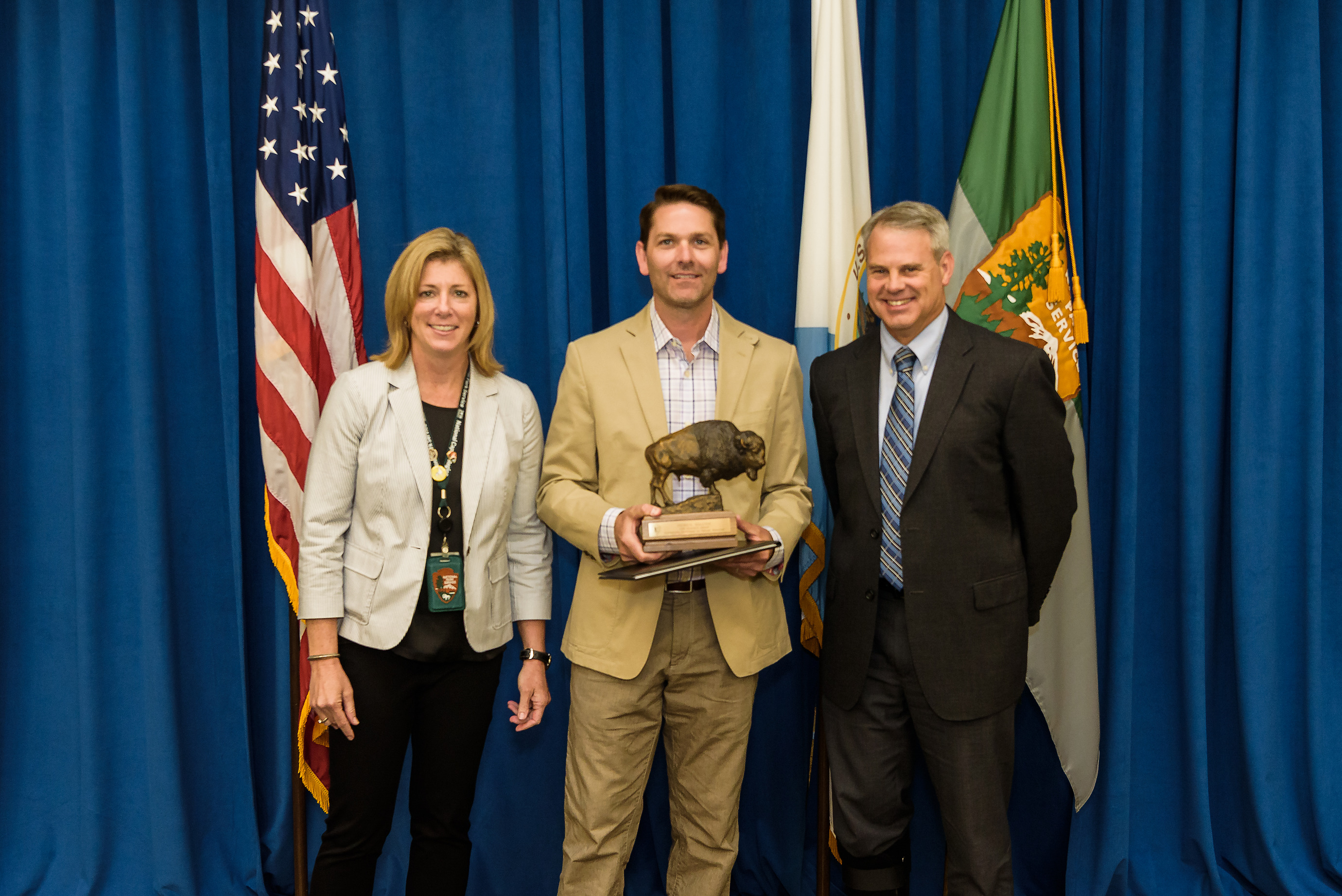 a man in khaki business suit holds a bison statue while standing in between a man and a woman. The background is a blue curtain with an American flag and National Park Service flag.