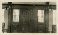This is an historic black and white photograph from the Scotty's Castle Historic Photograph Collection, Death Valley National Park featuring interior of front door entrance. Huge, textured, wood beam lintel across top. Side light each side protected by carved wood spindles. Paneled area below window. Central door is made of six massive boards held in place with handwrought metal rivets. Large, handwrought strap hinges top and bottom span width of door with short middle hinge. Unique handwrought door lock and latch.  Curtain rods at each upper corner. Tile and radiator grills at each lower corner.