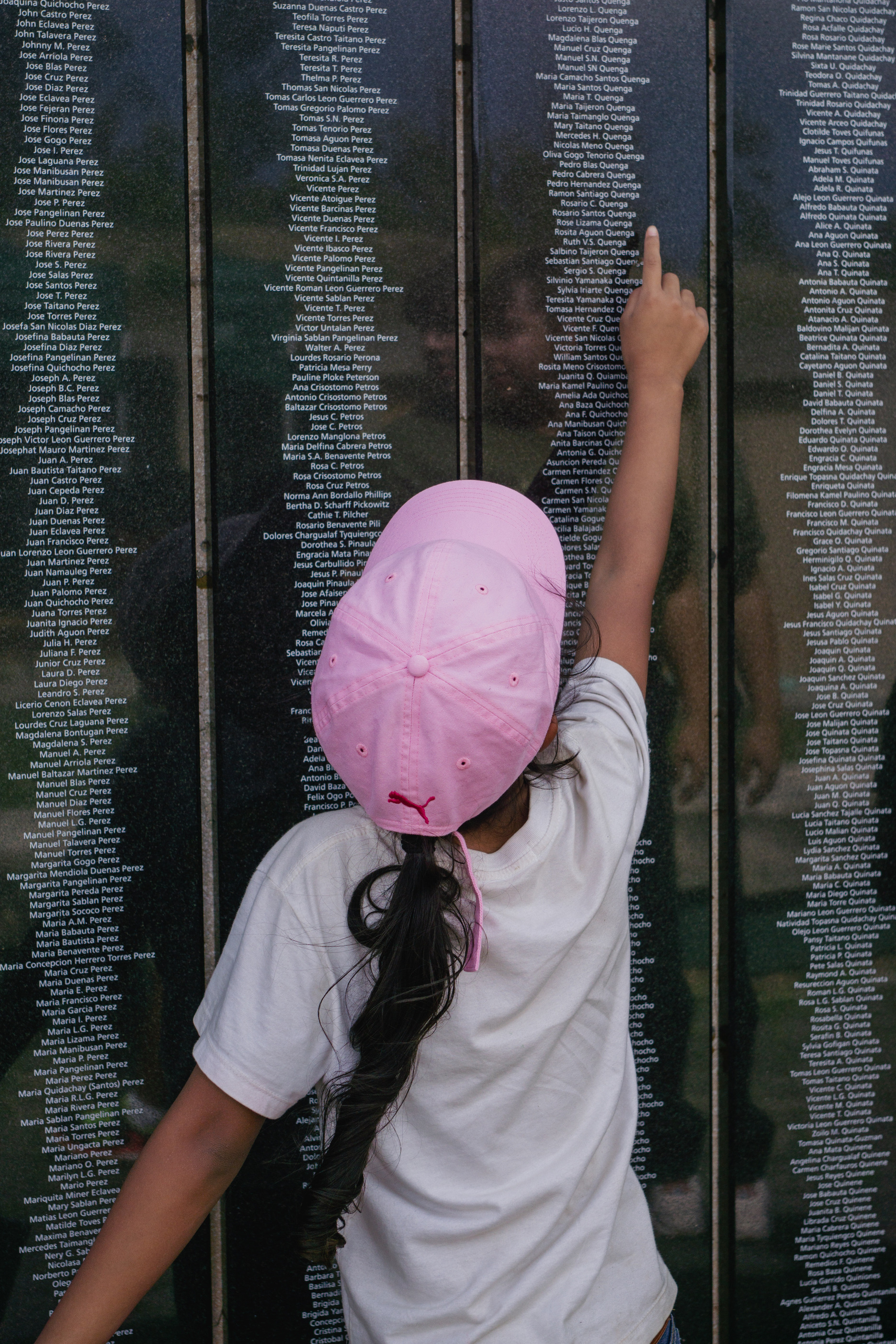 A little girl in a pink hat facing away from the camera points to the name Rose Lizama Quenga on a black plaque with names engraved on it.