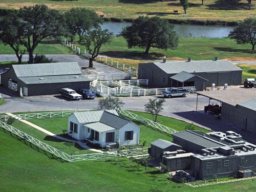 Aerial view. Three green buildings are surrounded by pavement. One white and one green building are surrounded by grass. White fences are around and in between the buildings.