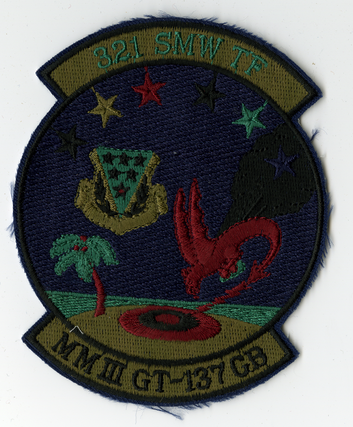 Dark blue circular with cut edges and with tab on top and bottom.  Top tab has "321 SMW TF" in spruce green on olive green. Bottom tab has "MMIII GT-147 GB" in black on olive green. Around top of circle are six (6) start, black, tan, red, black, spruce green, and black. Left center is 321st SMW symbol and along right is red dragon flying towards bottom left and tail curling under body. From rear is black plume of smoke and from tip of tail is red lightning bolt. On bottom of circle is olive green island with spruce green and red palm tree on left and multiple circles in center forming a target, red, black, with red bulls eye.
