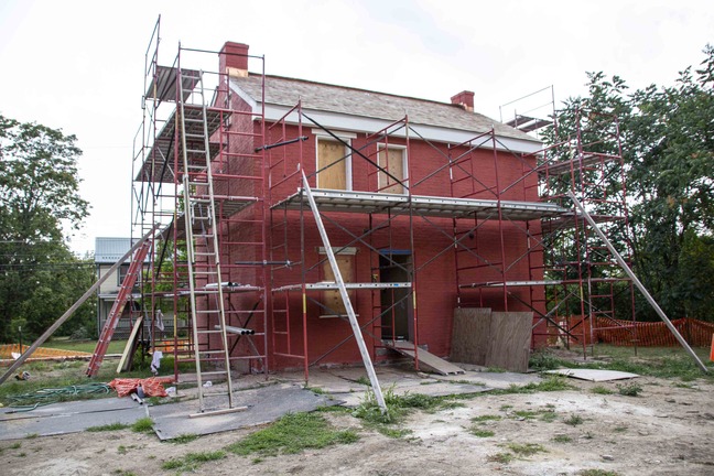 There is red scaffolding surrounding the house with an orange ladder propped up against the side of the house. A green water hose can be seen next to a pile of orange construction fencing. There are two wooden boards beside an open backdoor leading into the house. 