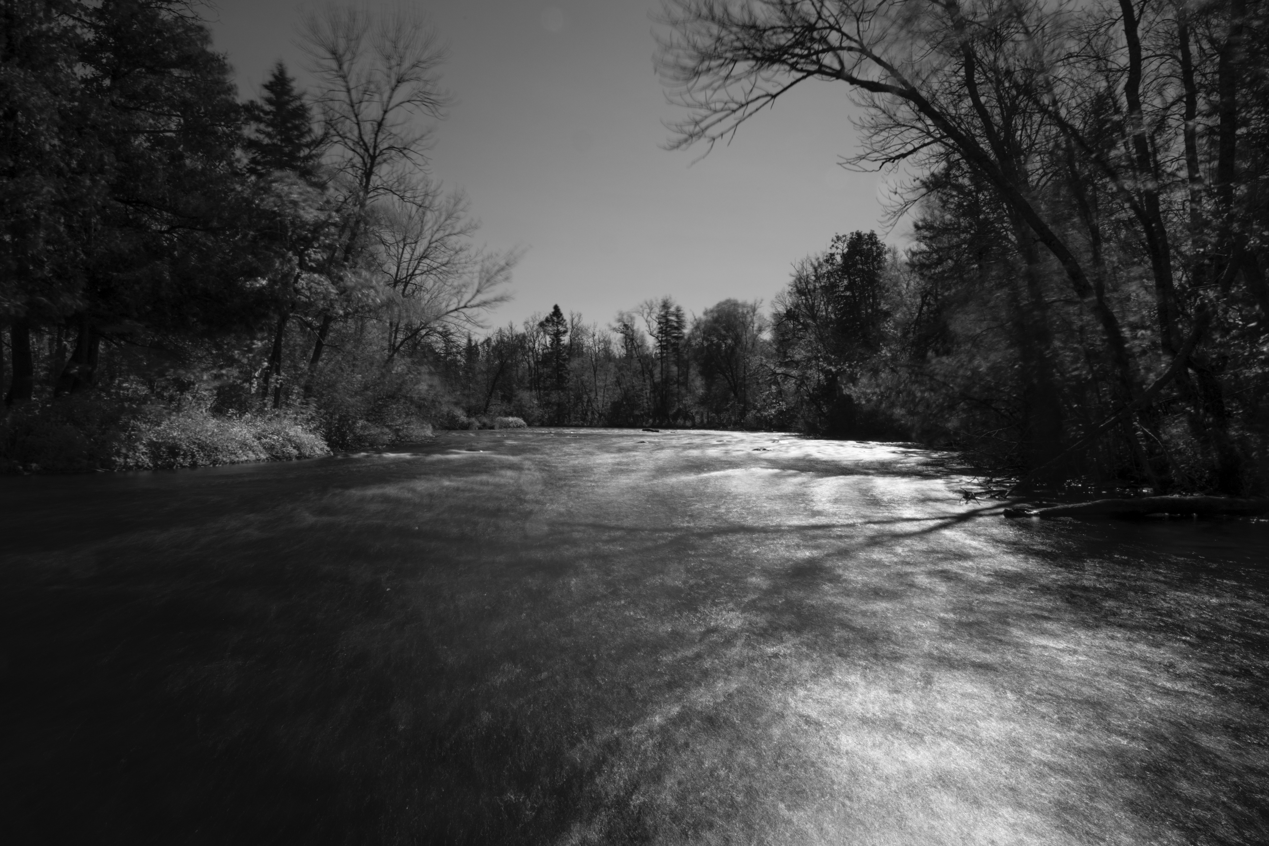 Though clear in subject, the image appears with the blur of motion indicative of a long exposure. Distal branches of trees blend into the clear sky beyond while a river rushing towards the photographer’s perspective is softened. It is because of this softness provided by long exposure that the shadow of a tree can be seen on the water’s surface – a detail that would be obscure to the naked eye.