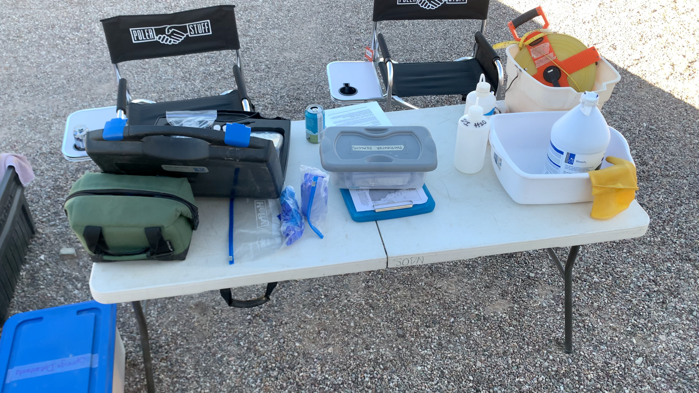Plastic folding table holds a variety of gear cases, boxes, and plastic bottles. A gallon jug and pair of rubber gloves sit in a plastic wash basin.