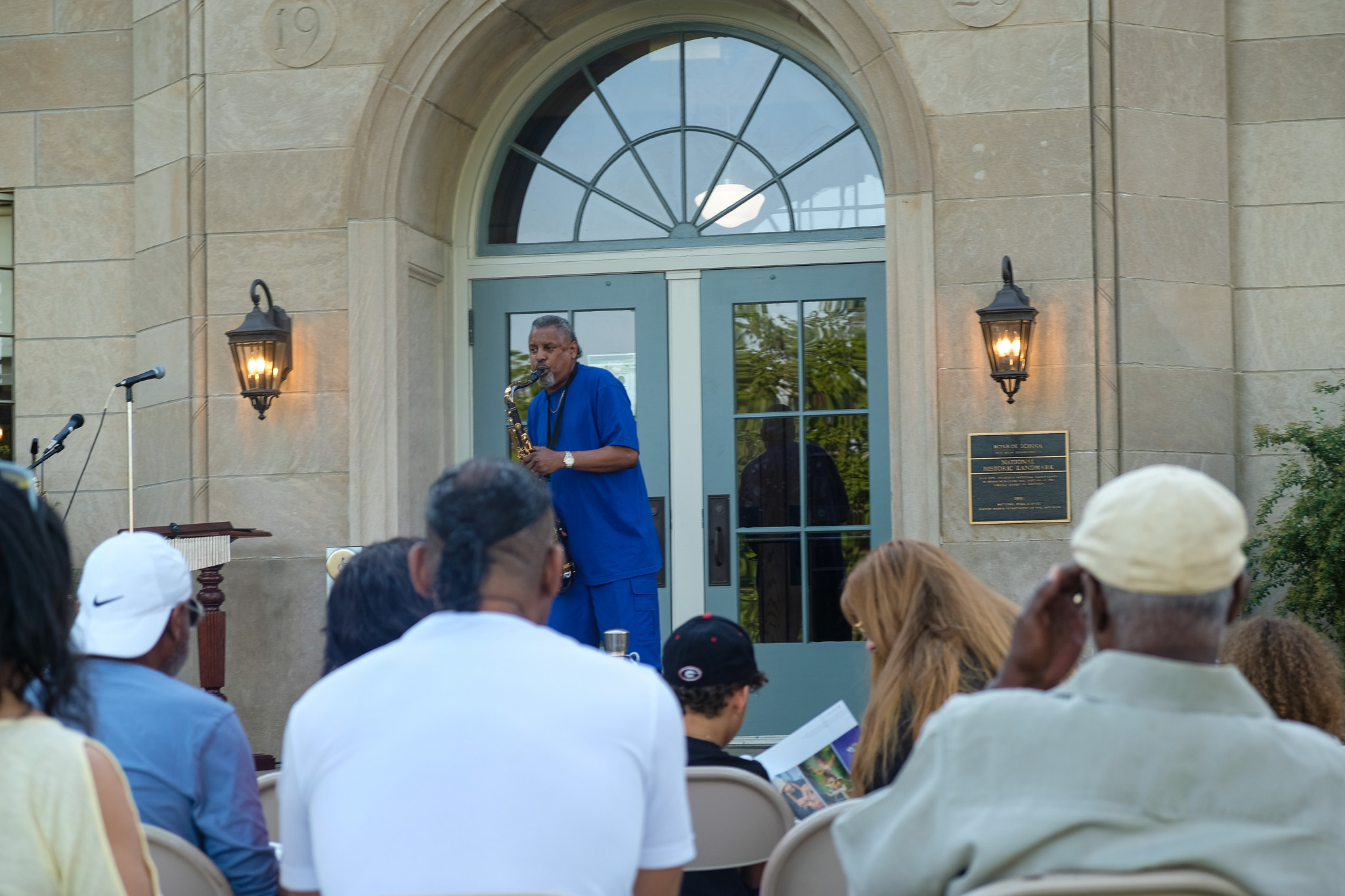 An African American man can be seen in the center of the screen playing a saxophone. A crowd of seated onlookers can be seen in the foreground. An early 20th century school made od sone and bricks is visible in the background.