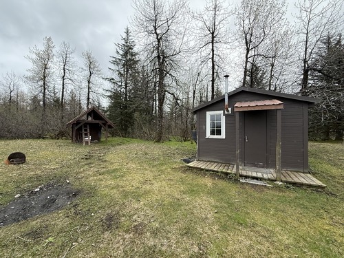 A small cabin and shed on a grassy lawn with trees behind it. 
