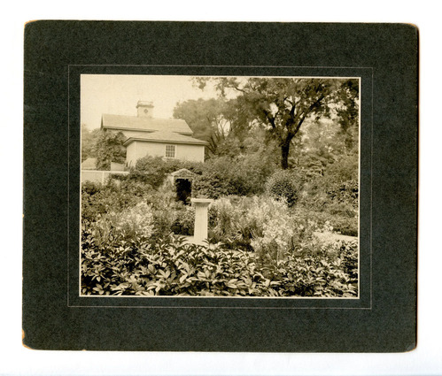 Black and white photograph of formal garden in full bloom with empty pillar in center.