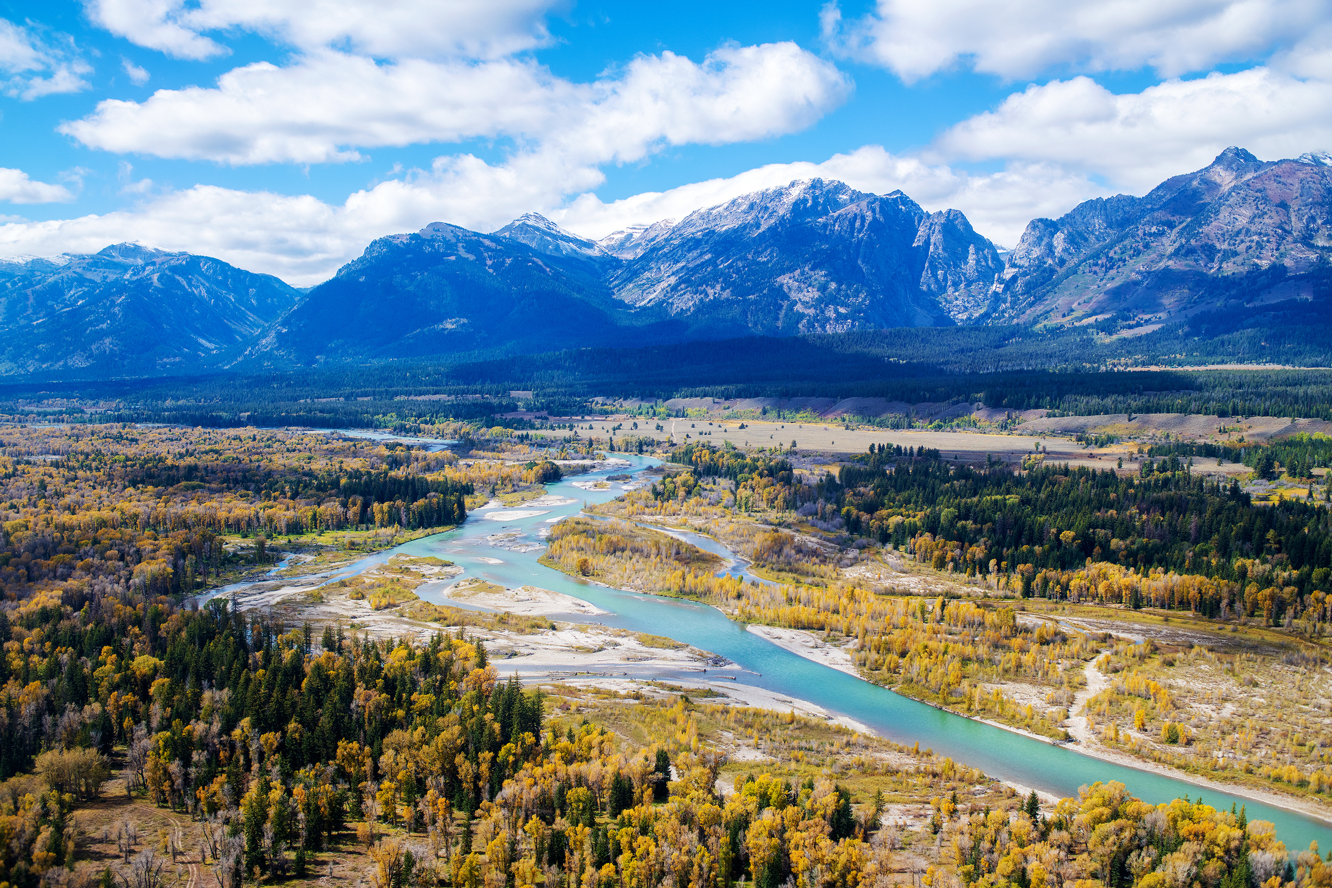 Arial view of aquamarine winding river, yellow and green trees, and cloud-shadowed mountains in the background. Vibrant colors throughout.
