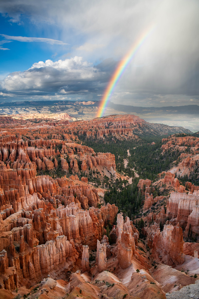 A vivid rainbow arcs down from a raincloud into a landscape of red and white rock spires and cliffs.
