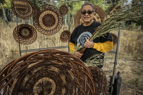A man holds willow cuttings while standing in a booth displaying handmade willow baskets and platters.