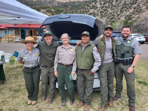 Six park rangers pose in front of an electric pickup truck at an outdoor community event.
