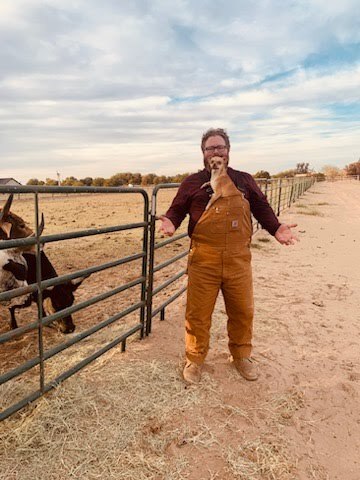 a young man dressed in tan overalls with a small dog peeking out the top, next to a gate with cattle