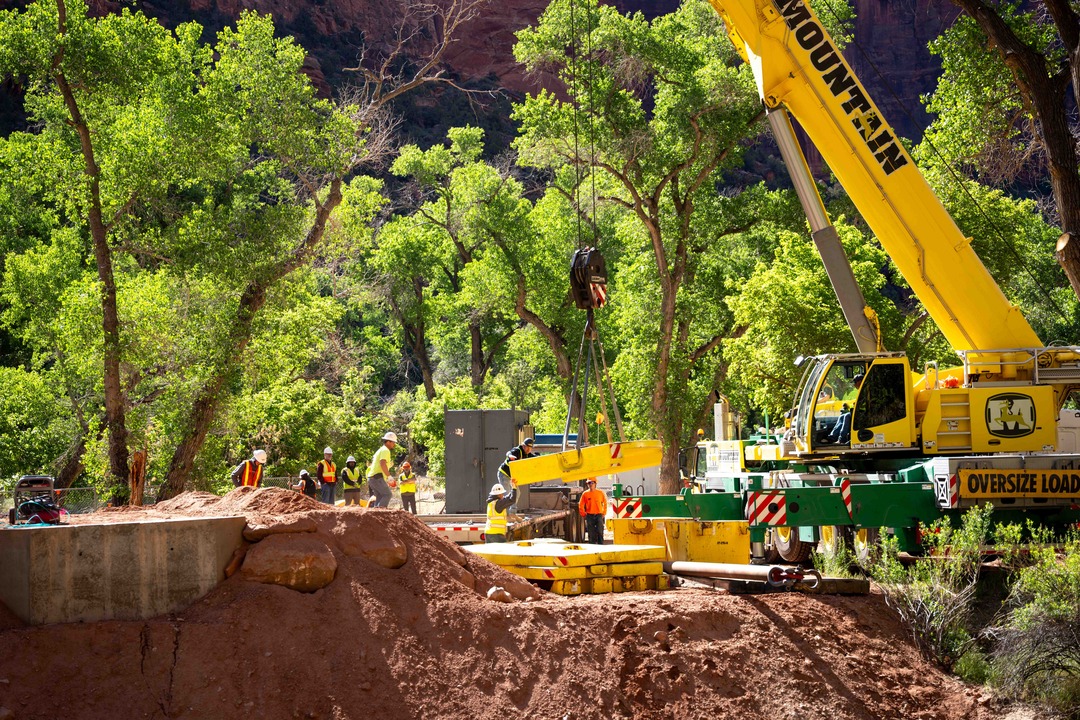 A giant yellow crane picking up large yellow structures with construction crews, trees and sandstone in the background