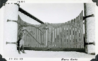 This is an historic black and white photograph from the Scotty's Castle Historic Photograph Collection, Death Valley National Park of man in cowboy hat, coat, vest, knee high boots looking at large gate with matching sides. Each side has vertical boards of various length to form curved top edge. Decorative wrought iron hinges are massive leading from stucco pillar to last board on gate.  Wrought iron gate closure at top. At bottom is hinged pin that can drop down to keep gate open. Stables tile roofline in background. Inscriptions in black ink along upper and lower border.