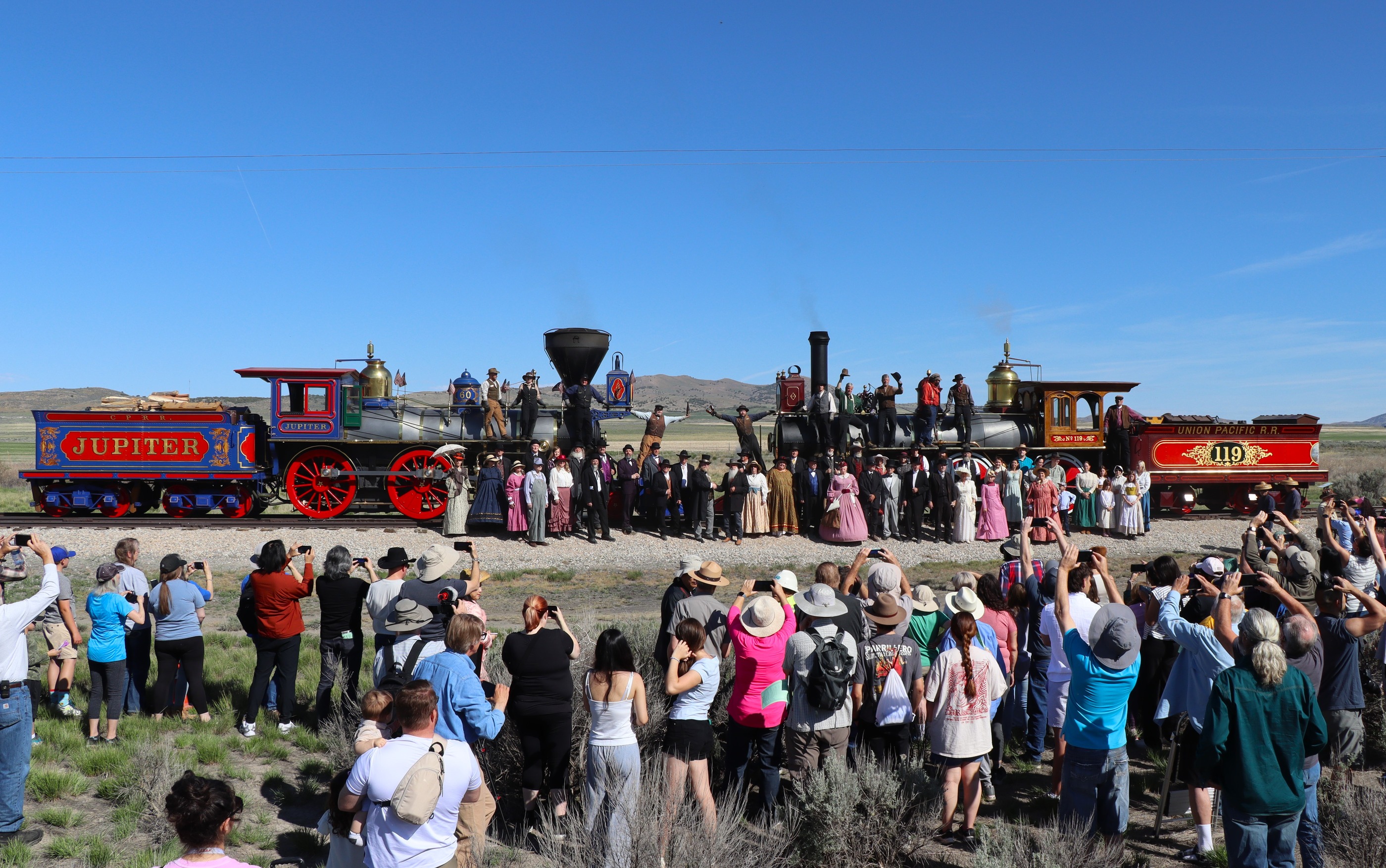 Men and women in turn-of-the-century costumes stand in front of two vibrant historic locomotives, while two engineers raise champagne bottles. Visitors in foreground taking photos