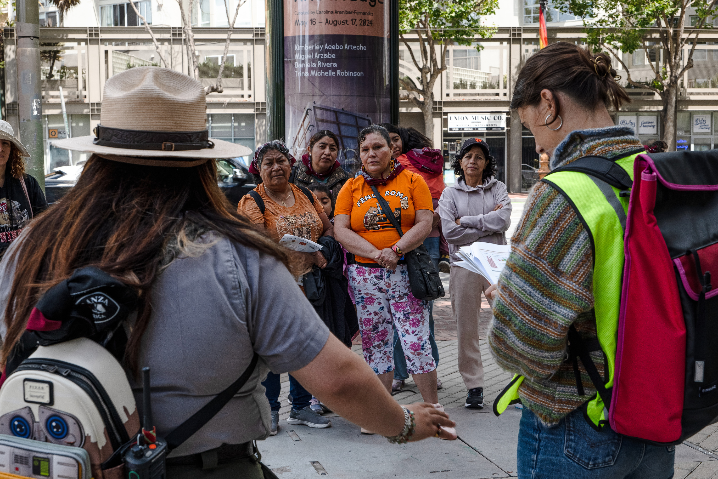 A group of women and children gather together on a city sidewalk to listen to a speaker wearing a high vis vest and an NPS Park Ranger