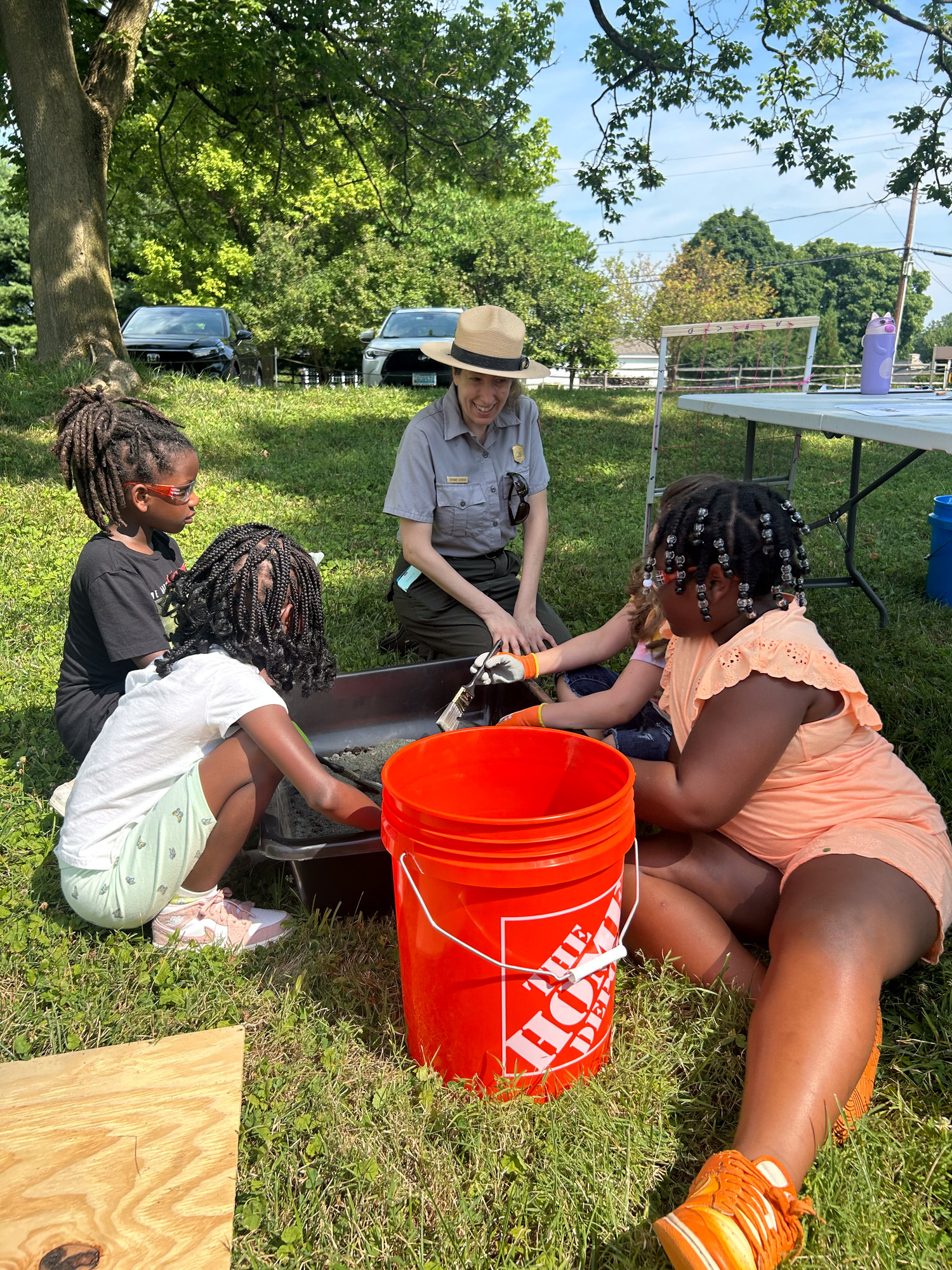 Park ranger with four smiling children. All of them are digging in the dirt using brushing and archaeological tools.