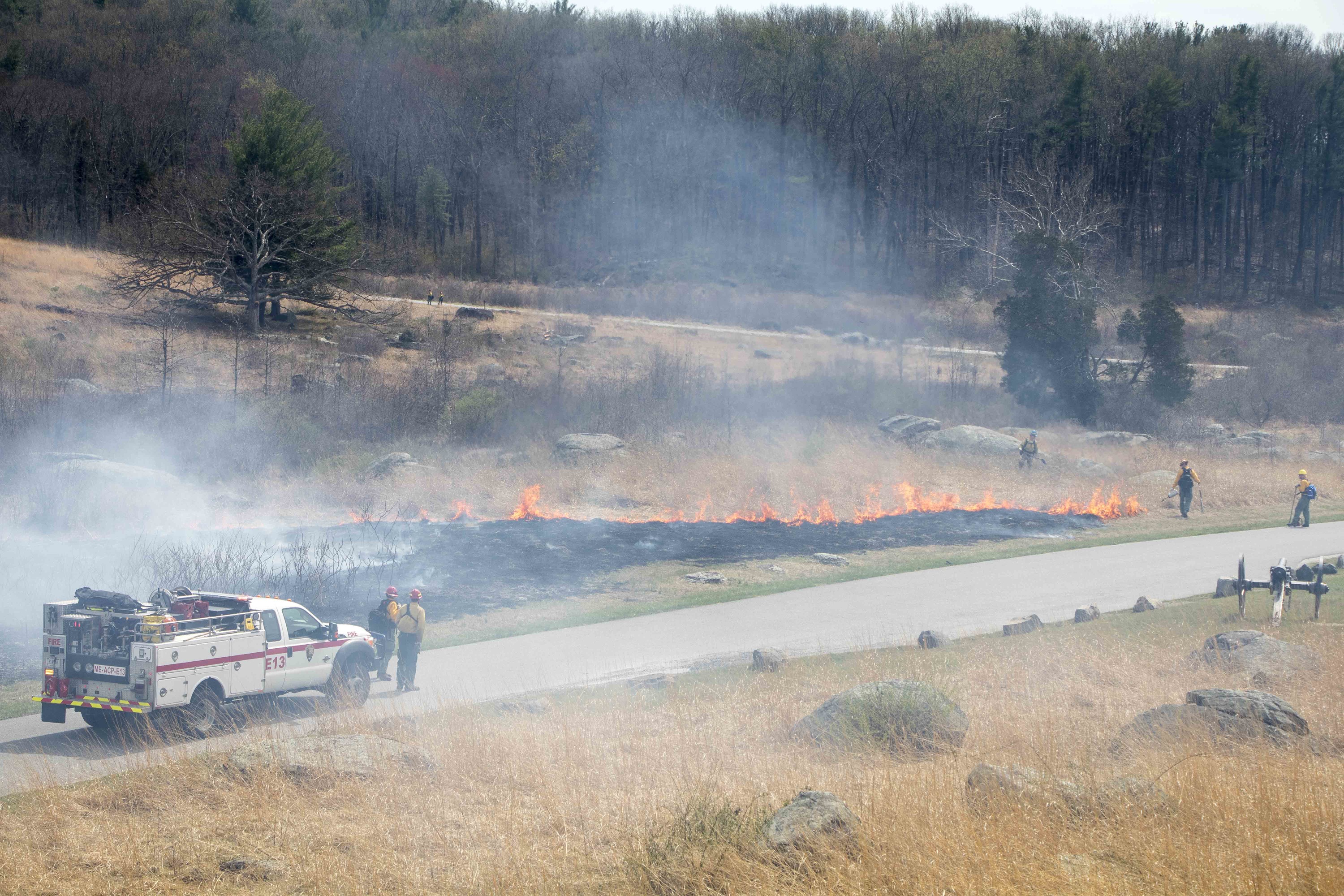 The prescribed fire moves away from Crawford Avenue. There is a fire truck in the road and four fire fighters monitor the area. The fire is moving away from the road.