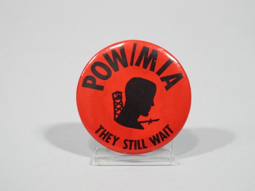 This image shows a round button pin with a red background and black text around the edges that says "POW/MIA" and "THEY STILL WAIT". In the center is a silhoutte of a prisoner.