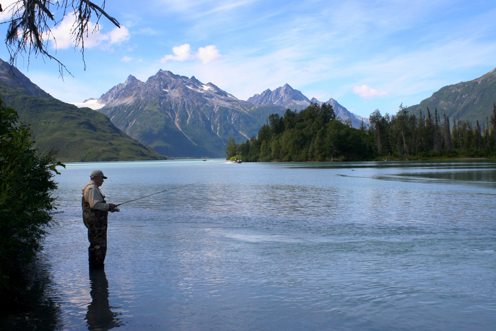 A fisherman in waders stands in a river in front of distant mountains