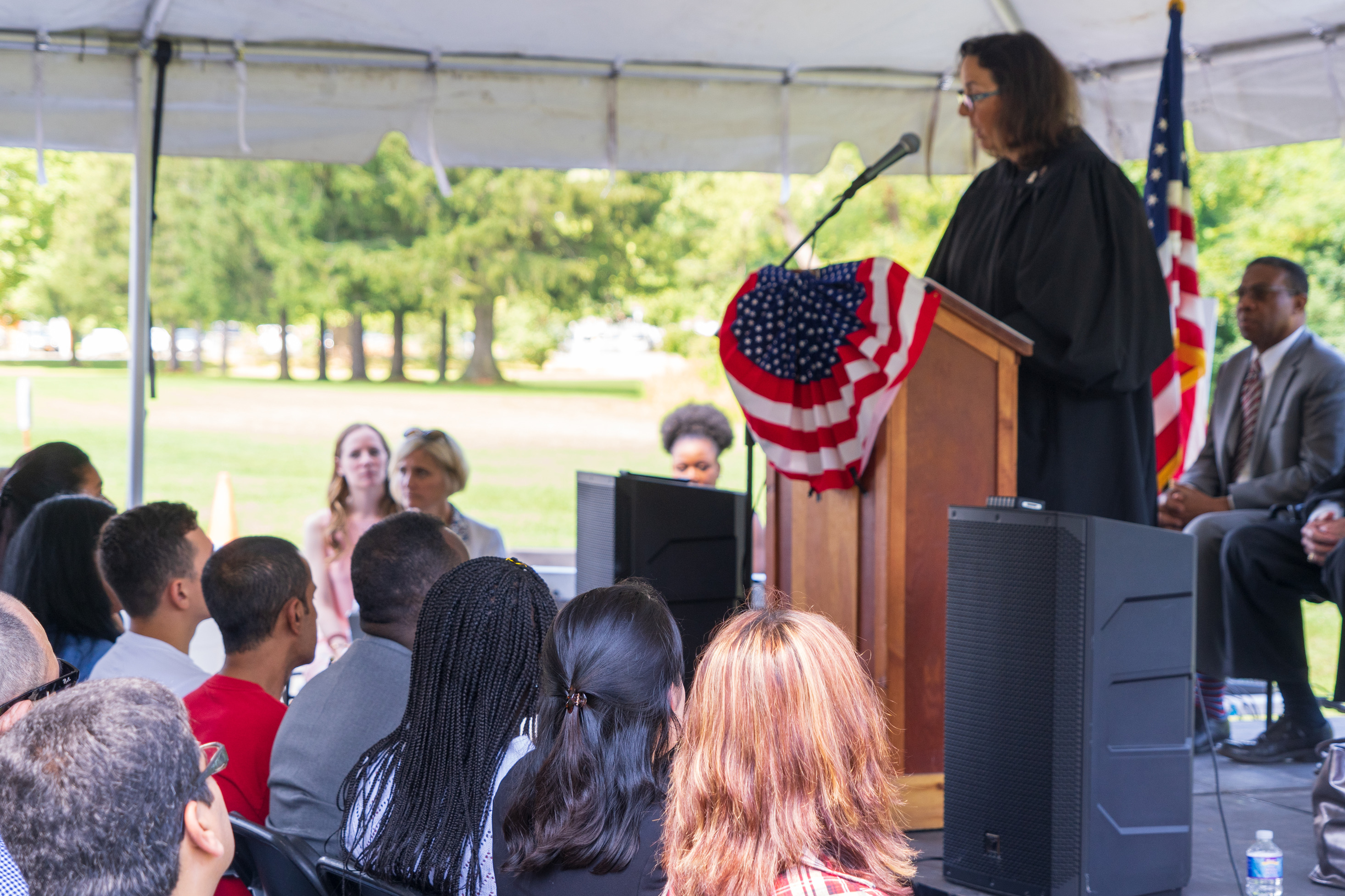 An audience sits under a tent and listens to a judge speak at a podium.