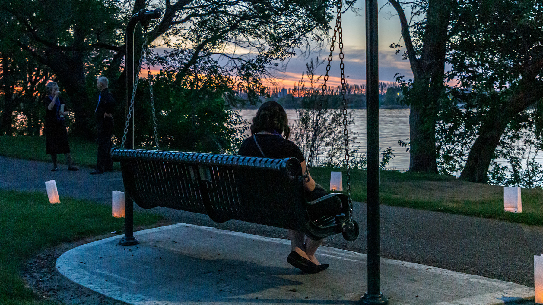 A person sits in front of a luminaria-lined pathway on a swinging bench that faces a river at sunset. 