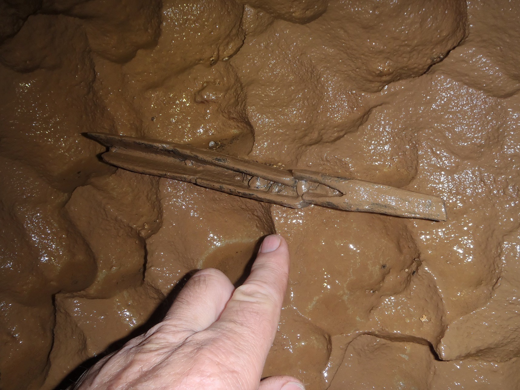A hand points to a small black spike covered in brown mud.