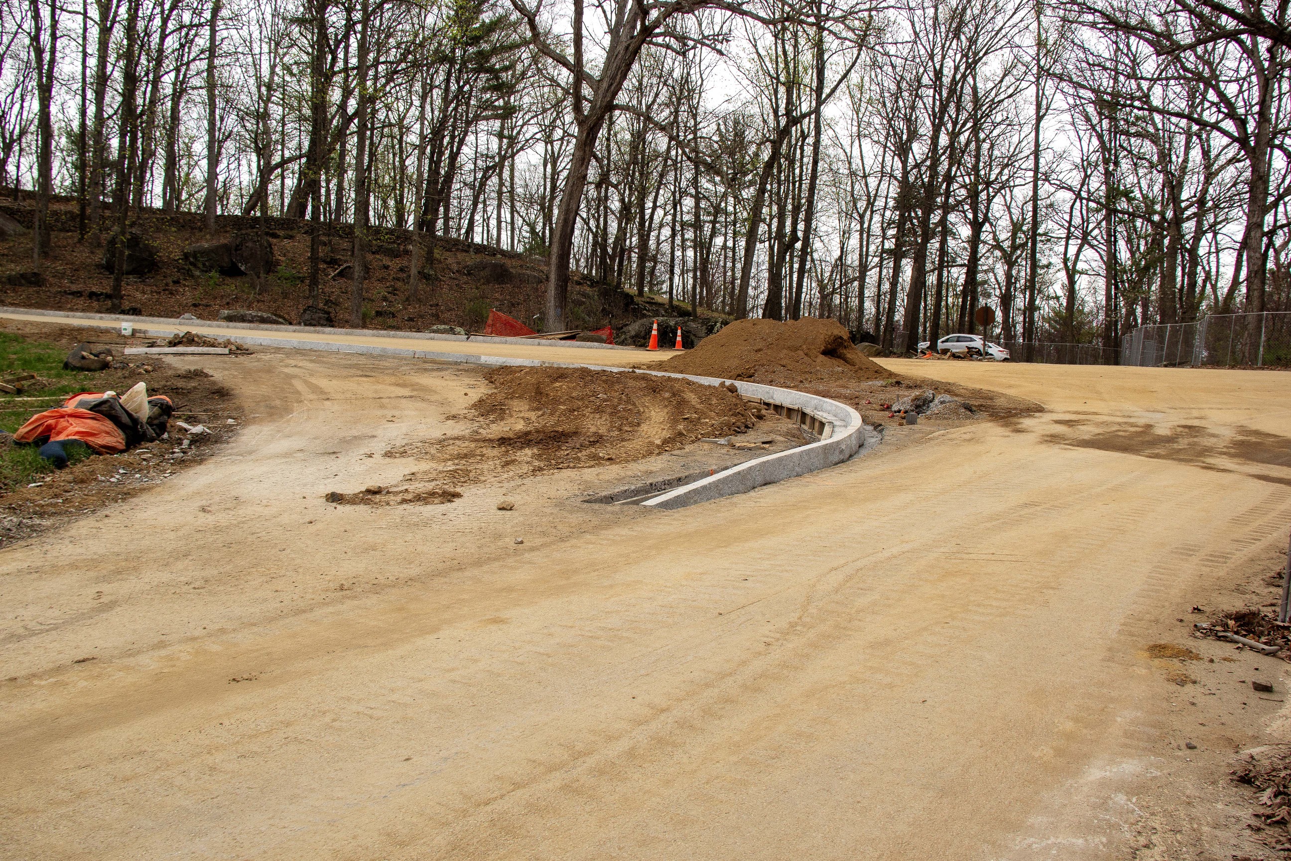 A dirt covered road with two piles of dirt on each side of a granite curb. In the upper left there is a line of trees. In the right of the photo there are two white cars. 