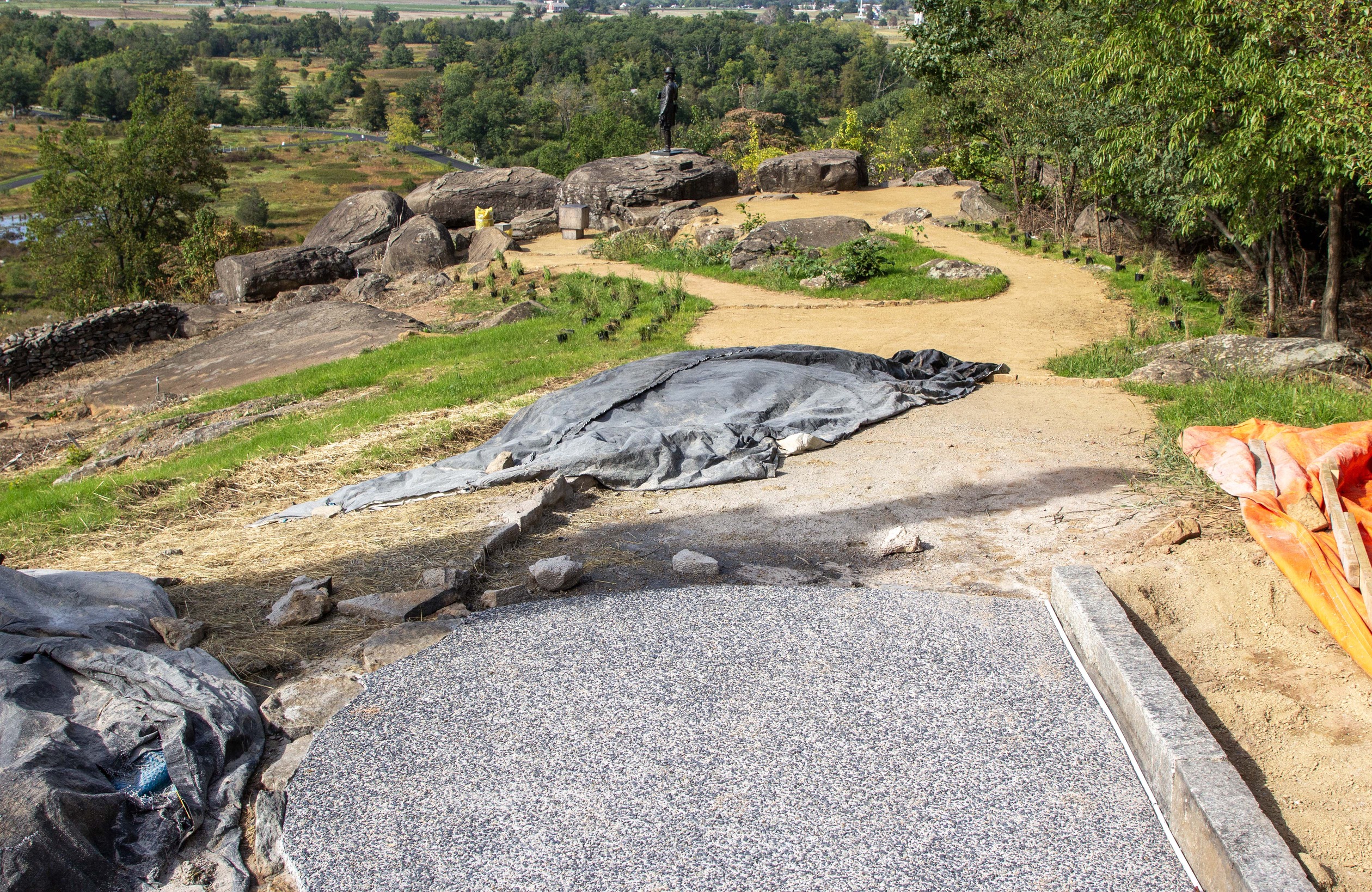 A concrete sidewalk ends near the bottom of the photograph. Beyond the concrete sidewalk, the trail transitions to a light brown colored gravel trail. There are multiple black tarps in the center and to the left while a bright orange tarp is on the right. Many large boulders are in the distance and the gravel trail goes around many of these boulders.