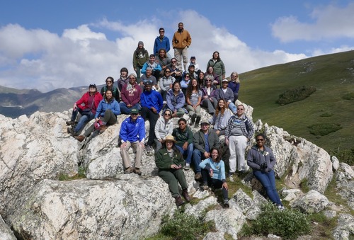 A group of 20+ interns pose for a photo on a outcropping of white rock with green tundra vegetation in the background. 