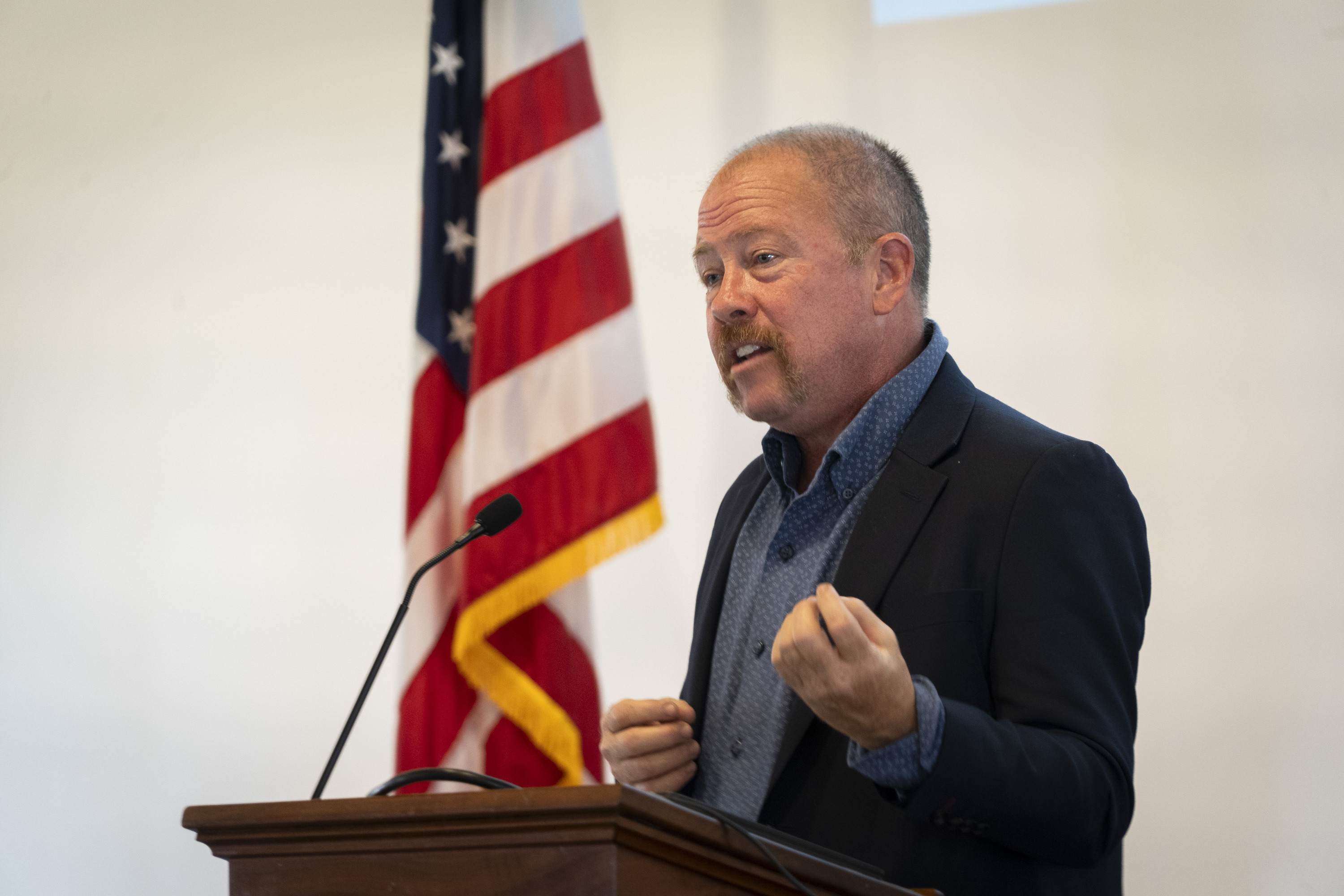 A man stands at a podium in front of an American Flag.